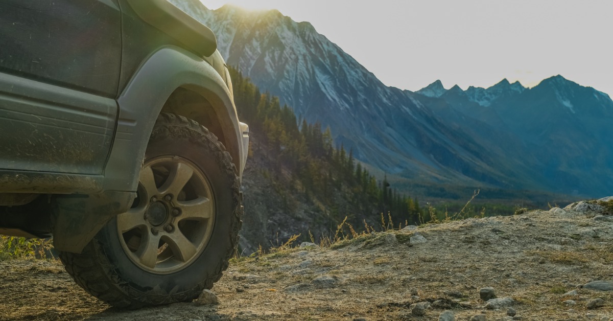 A close-up of a vehicle's front off-road tire on top of rugged terrain. A large mountain looms in the background.