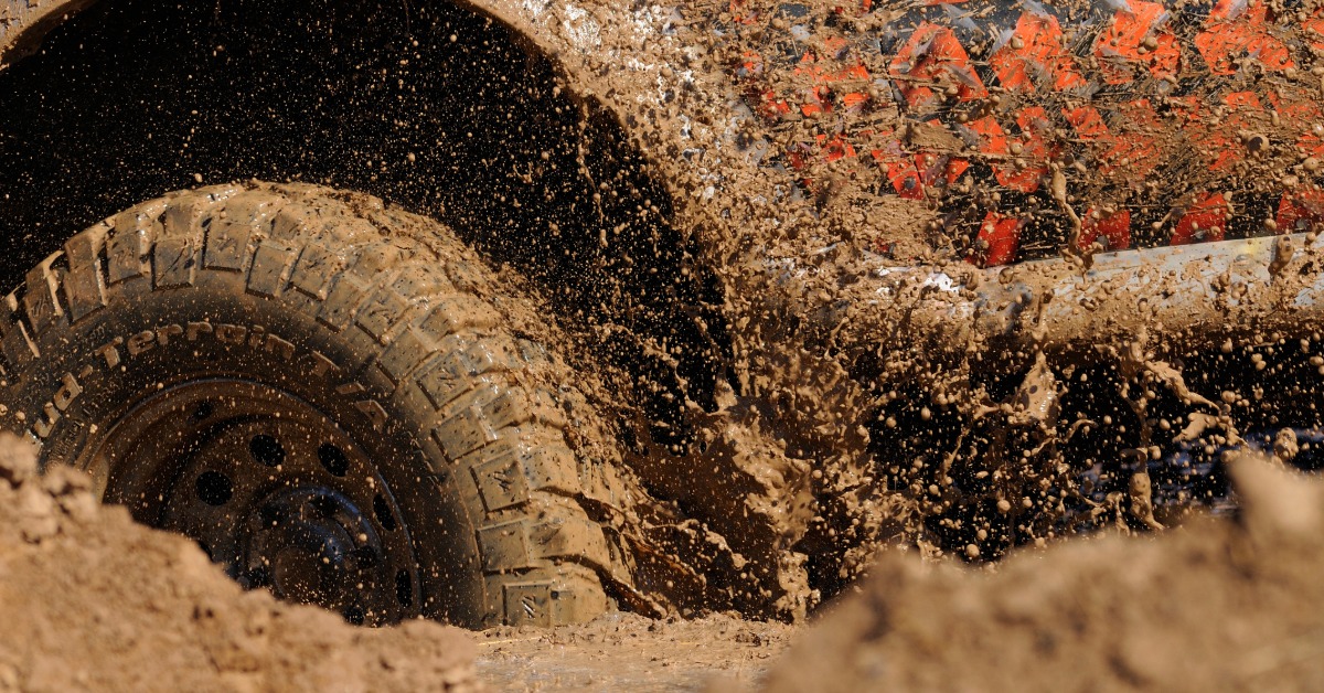 A close-up of a tire submerged in a mud puddle. Mud flies out of the tire's tread and sticks to the side of the vehicle.