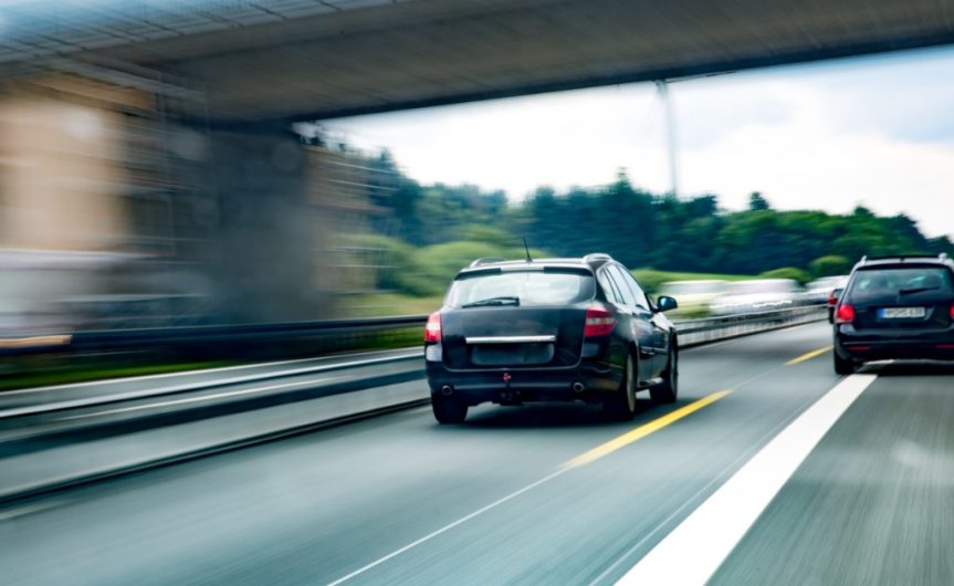 A black car traveling on a multi-lane highway under an overpass with other vehicles and blurred road lines.