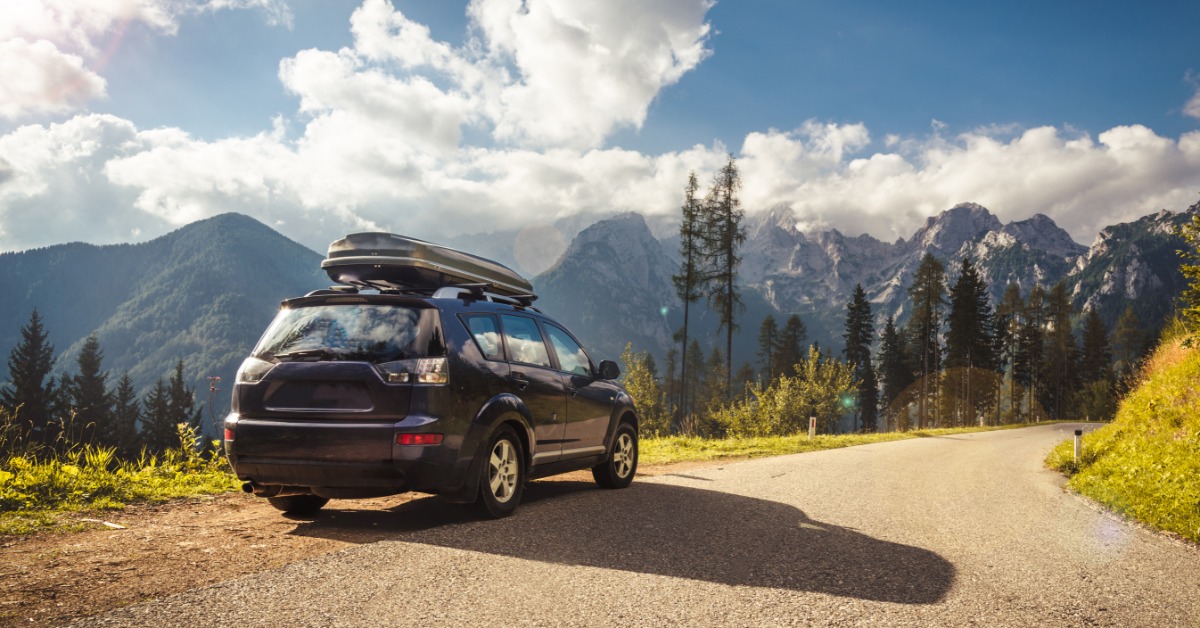 A dark SUV with a roof cargo box is parked beside a winding mountain road with trees, clouds, and rocky peaks.