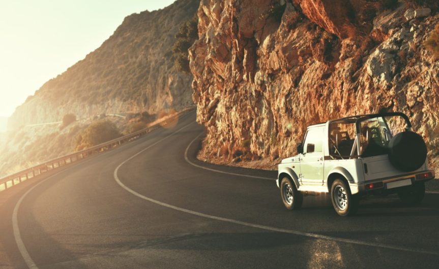 An SUV driving on a winding country road near rocky hills and water beneath a warm sunset sky with mountains behind it.