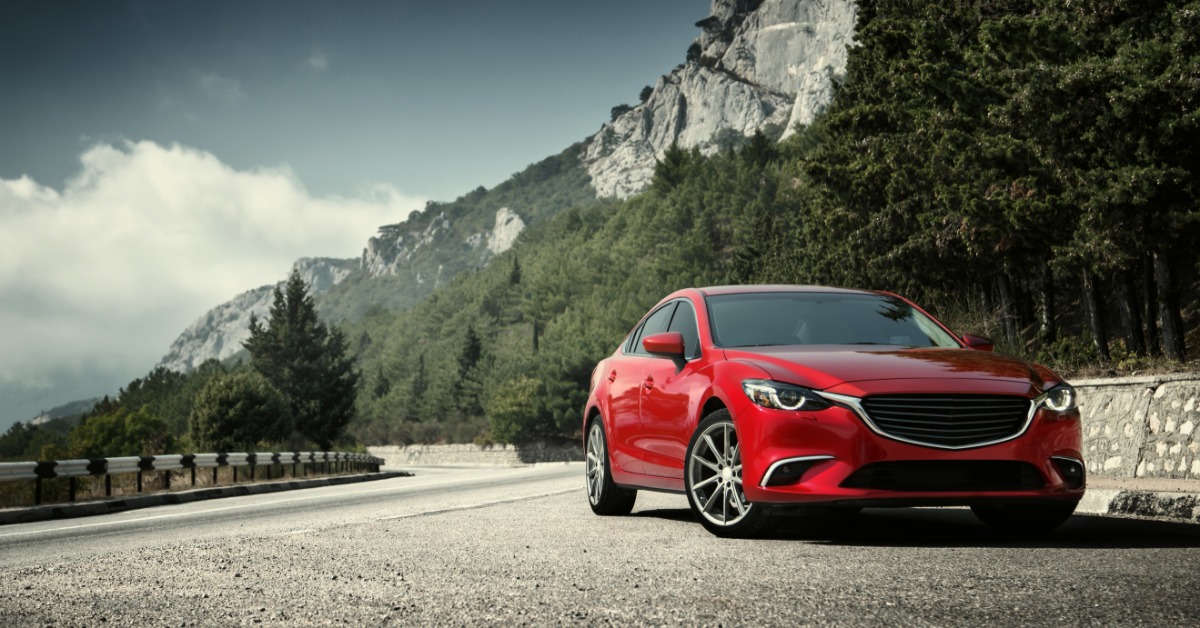 A red car parked on a paved mountain road with green hills, trees, rocky peaks, and a cloudy sky in the background.
