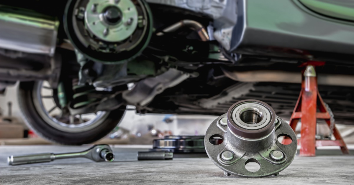 A wheel bearing rests on the floor of an automotive shop. A car is on a jack in the background with a wheel removed.