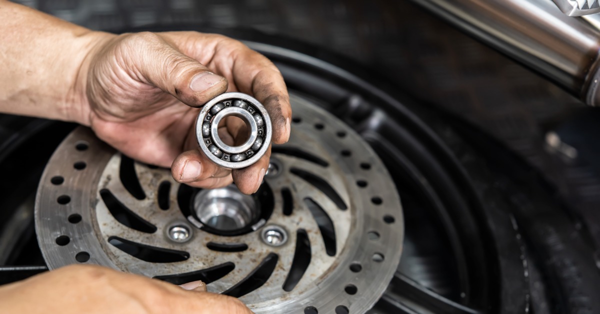 Someone with greasy hands holds up a small ball bearing above a brake rotor. A black wheel and chrome exhaust sit behind it.