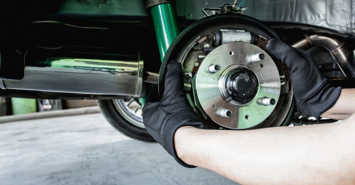 Someone wearing black gloves holds onto a wheel bearing hub still attached to a vehicle. An exhaust pipe is visible.