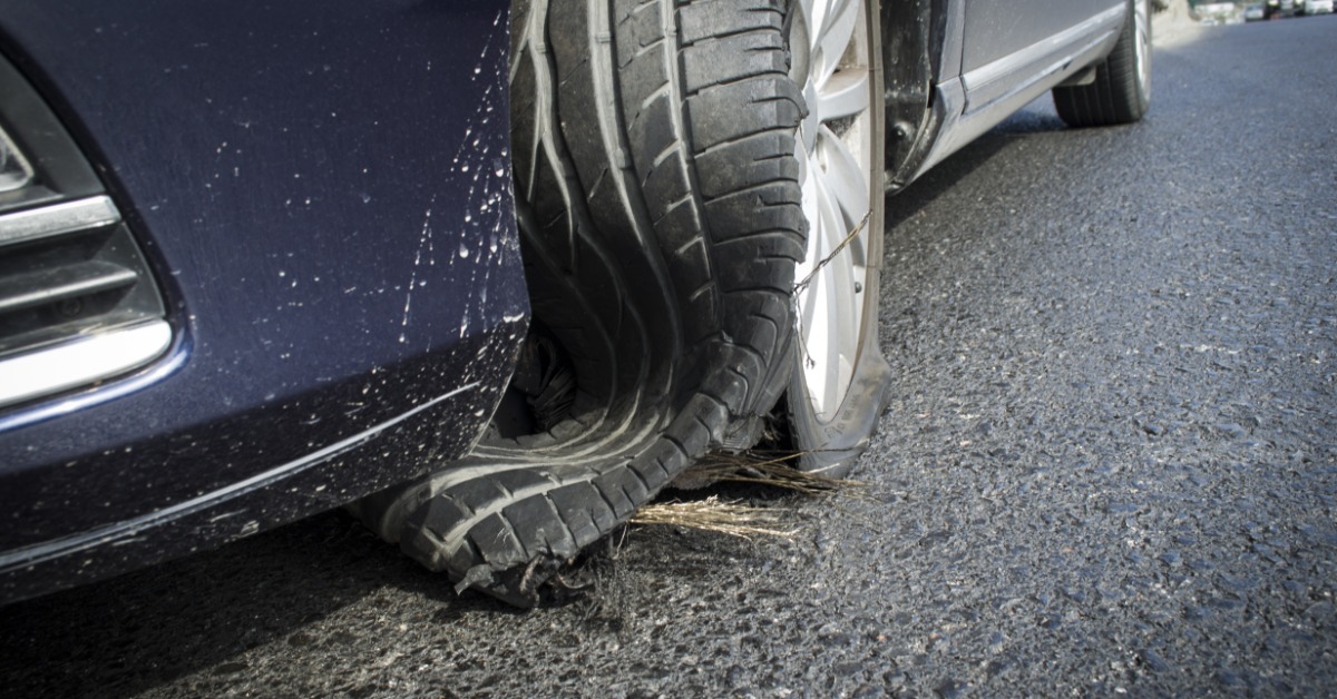 A close-up of the front wheel of a blue car on pavement with the tire blown out, with cars in the background.
