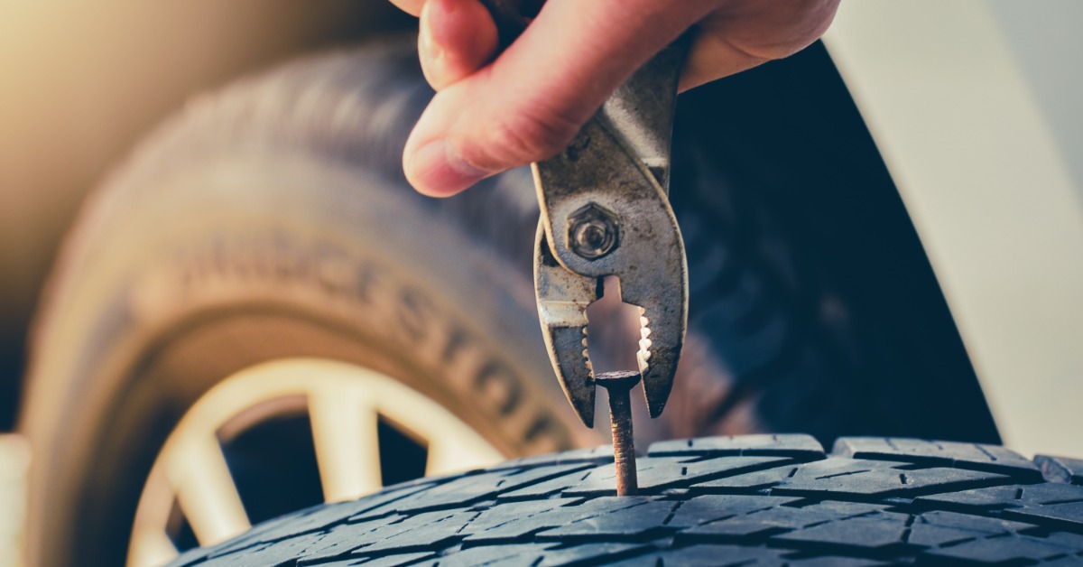 A close-up of a hand using a pair of pliers to remove a rusty nail from the tread of an automotive tire.