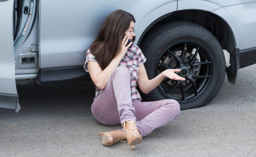 A woman is sitting on the road, leaning against her car, and talking on the phone with a flat tire next to her.