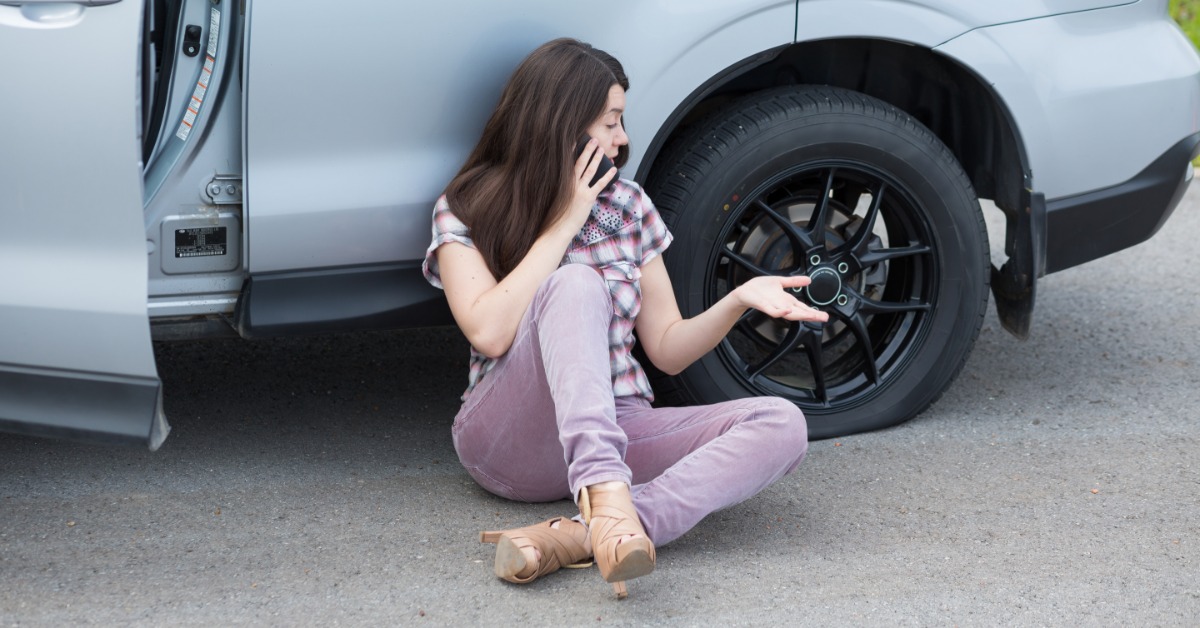 A woman is sitting on the road, leaning against her car, and talking on the phone with a flat tire next to her.