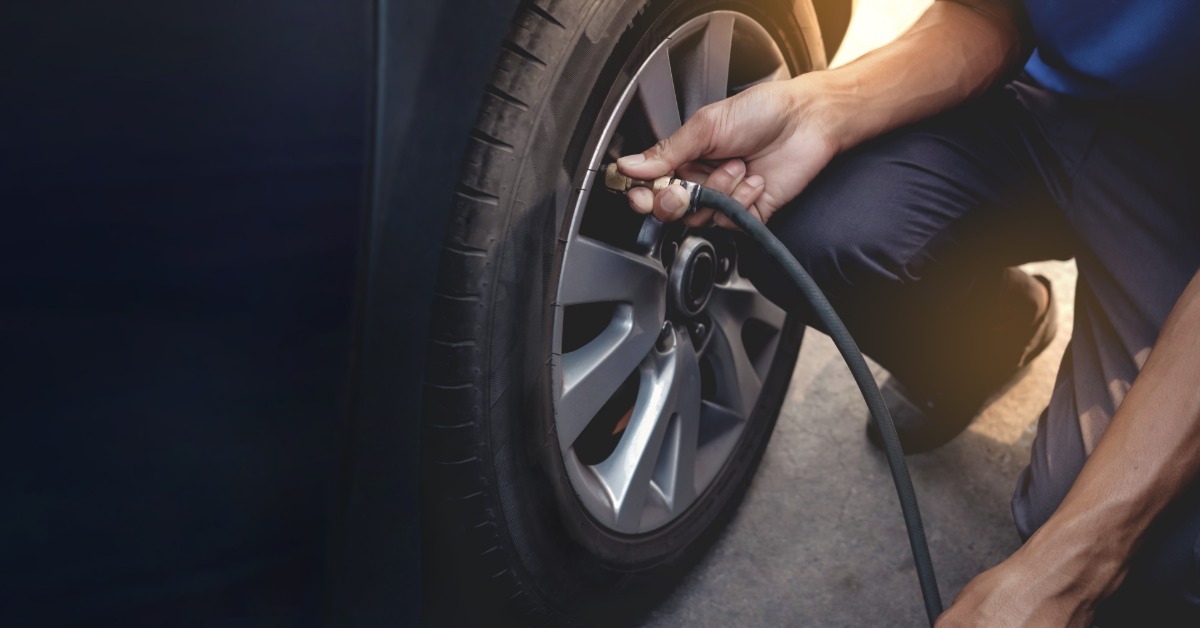 A close-up of someone kneeling next to a vehicle's rear tire. They hold an air hose up to the tire valve.