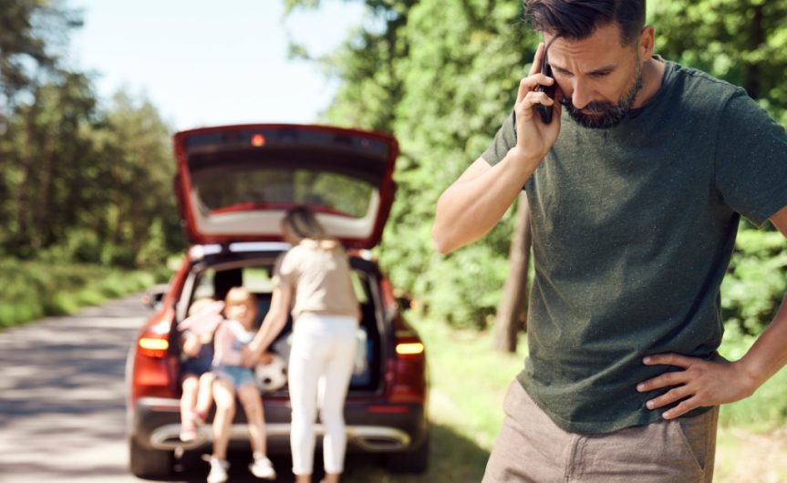 A man using a phone beside a red car with an open hatch, his family sitting near the back, and a warning triangle.