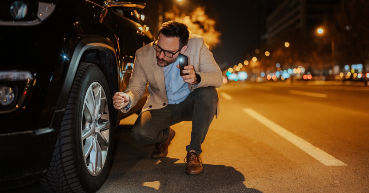 A man crouching beside a car tire at night while holding a phone flashlight along a dark urban roadside.