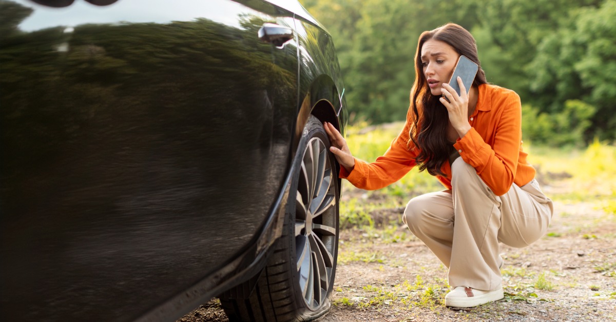 A woman crouching beside a black car tire while holding a phone near a grassy roadside with trees behind.