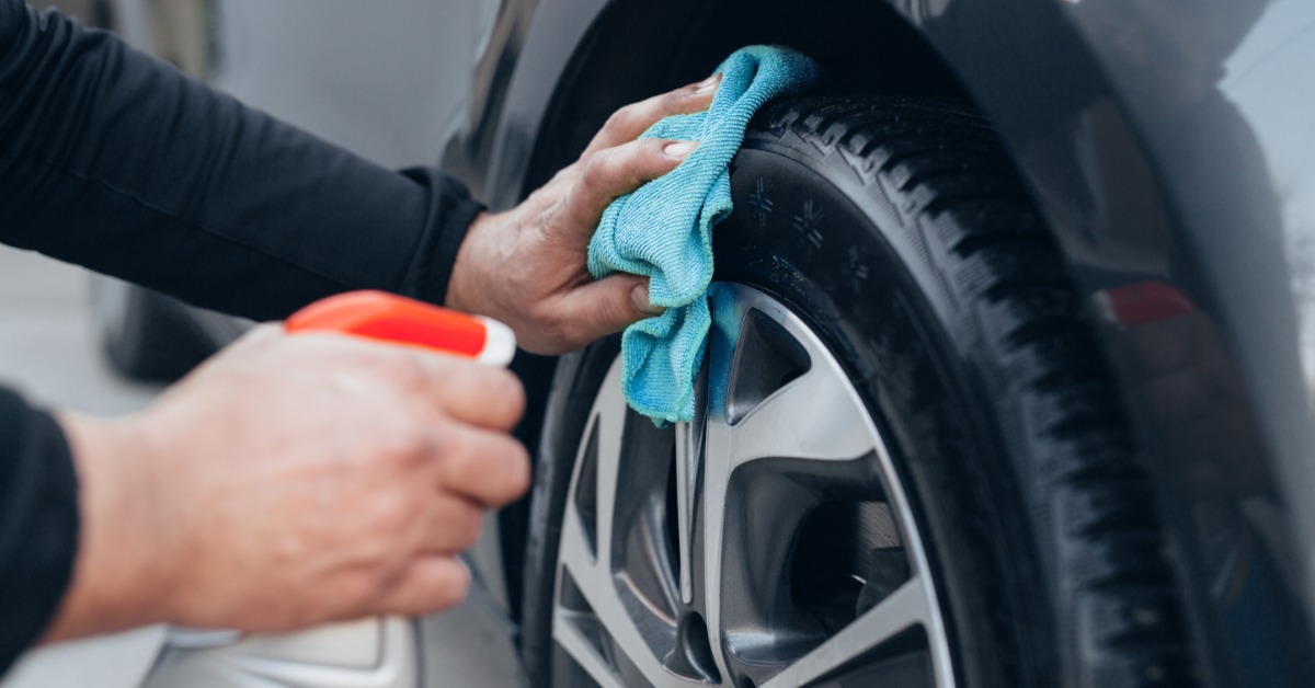 A close-up of someone wiping the tire of a car with a blue rag while the other hand holds a spray bottle.