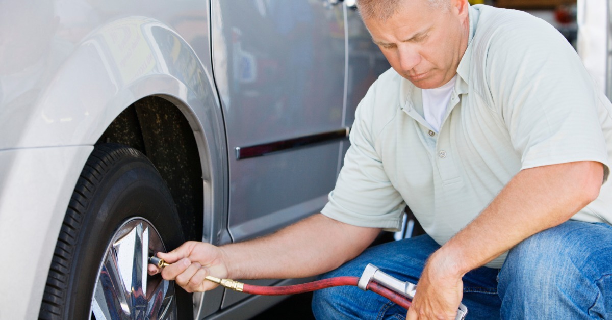 A man in a polo and jeans crouches down to place the nozzle of an air pump up to the tire valve stem of a car.
