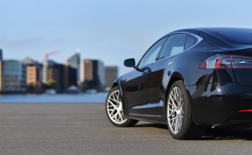 A rear view of a black luxury, four-door car parked on a city embankment with high-rise buildings in the background.