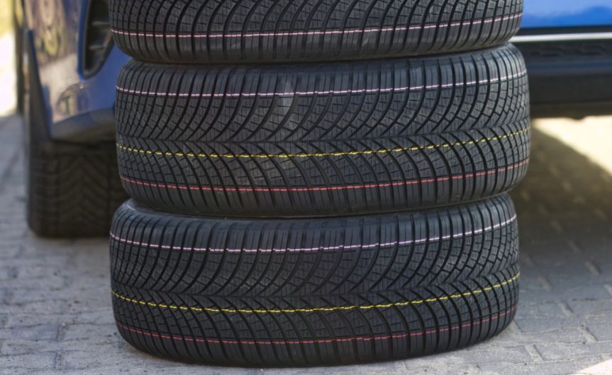 A stack of three new tires resting on top of each other. A blue vehicle is parked in a driveway in front of them.