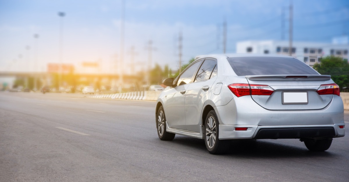 A small silver car drives down a highway with many lanes. Buildings and roadside greenery are blurred in the background.