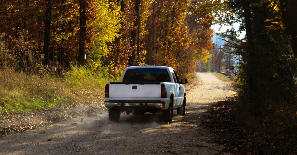 A white pickup truck drives over a gravel road in a wooded area. The trees' leaves are changing colors.