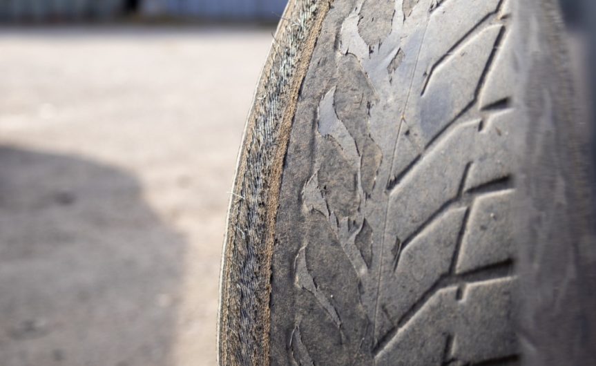 A worn car tire with shallow tread and exposed fibers on half the tire. It is resting on pavement beside a vehicle.