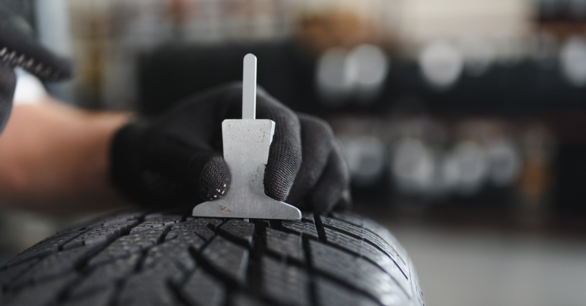 A gloved hand holding a metal tread depth gauge against a black tire with a blurred auto service area behind it.