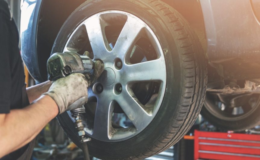 A close-up of a man with gloves using an impact wrench to unscrew the lug nuts on a car tire elevated on a lift.