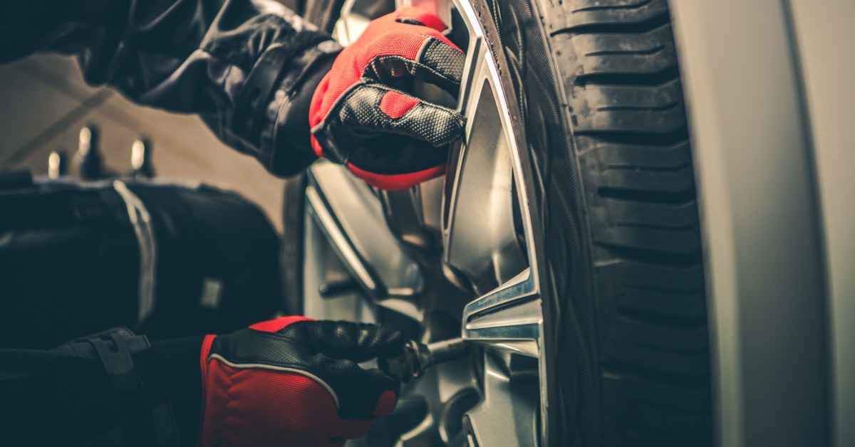 A close-up of a pair of hands with red and black gloves using a tool to unscrew the lug nuts of a car wheel.