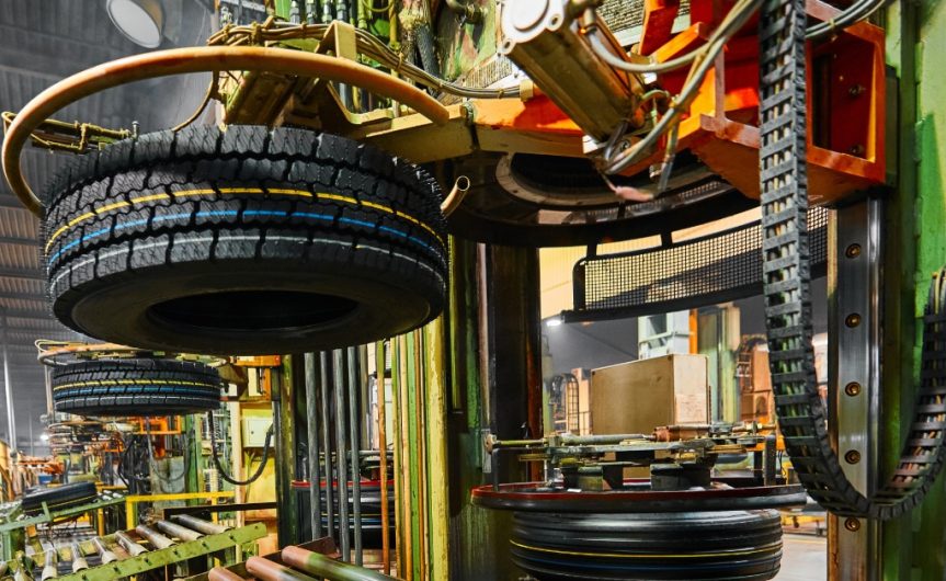 A close-up of a machine at a tire manufacturing facility holding up a brand-new tire above a roller conveyor line.