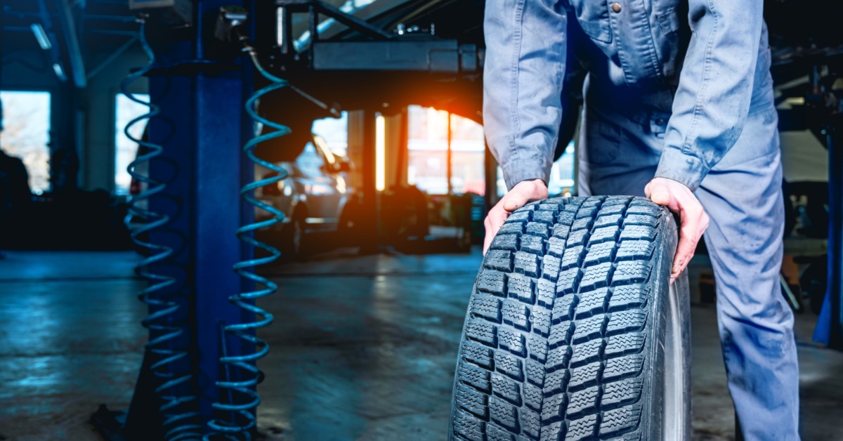 A close-up of a winter car tire freshly removed from a car being pushed by a man in blue overalls in an auto shop.