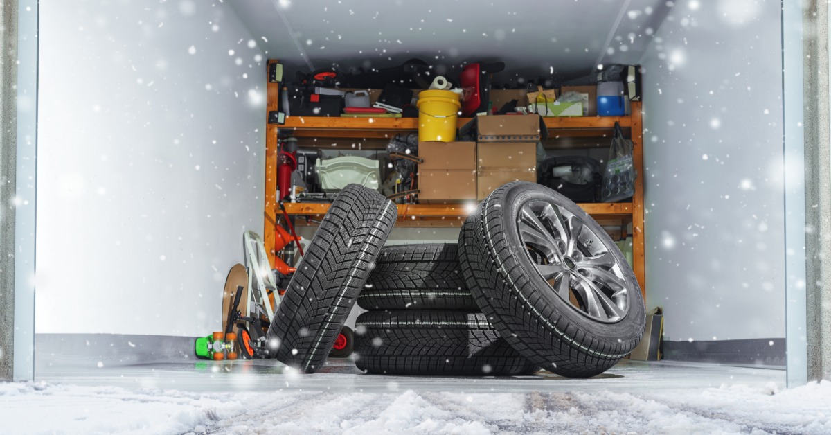 A set of winter car tires piled together in front of the opening of a storage unit with shelves and supplies behind them.