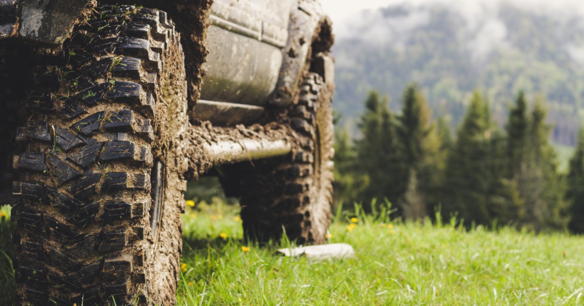 A ground-level close-up of an off-road vehicle with tires caked with mud on a grassy clearing with trees in the background.