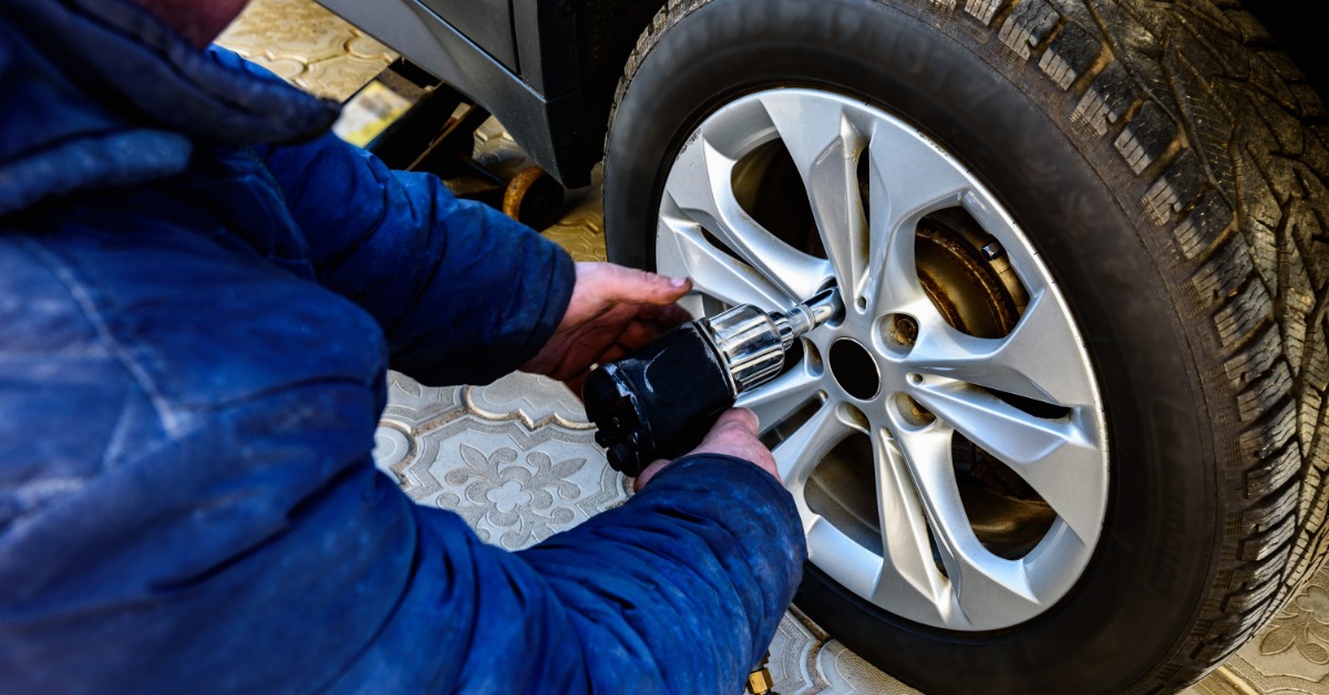 A close-up of a man in a blue jacket using a tire impact wrench to unscrew the lug nuts of an off-road tire.