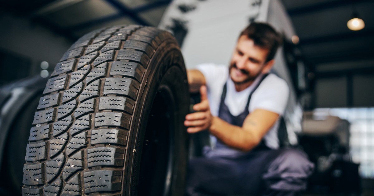 Mechanic crouching to pick up a brand-new tire with healthy treads in a large, brightly lit tire shop.