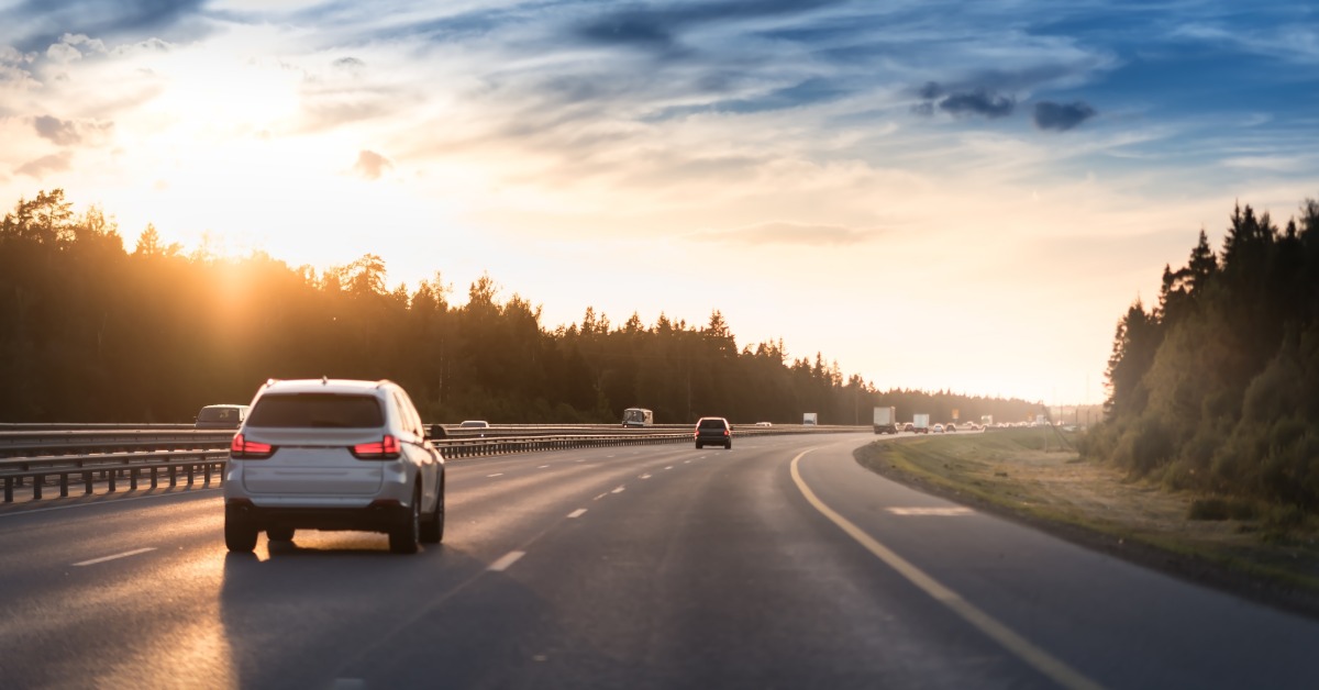 Cars on a highway with light traffic. The sky is a mix of orange and blue as the sun sets in the distance.