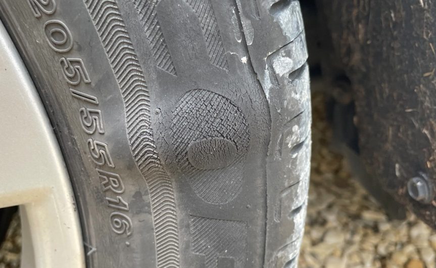 A car parked outside during the daytime. One of the tires on the vehicle is severely warped and dirty.
