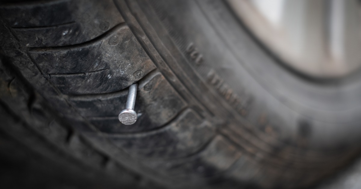 A long silver nail sticking out of a black, rubber car tire. The vehicle is parked in a brightly lit space.
