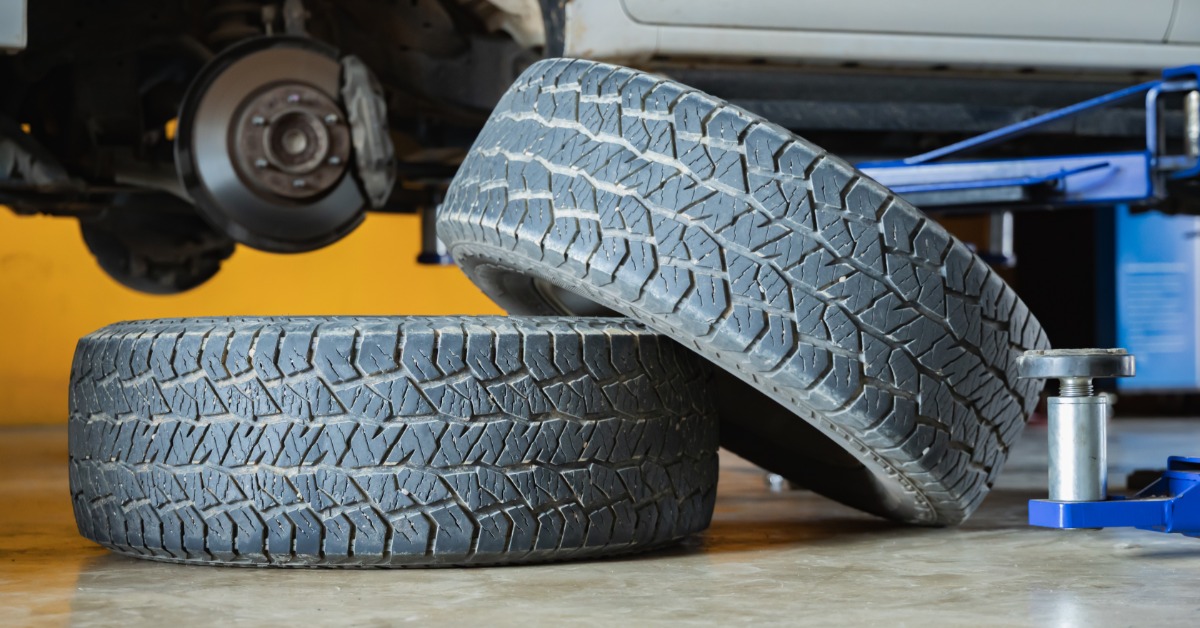 Two worn used tires on the floor of an auto repair shop. A vehicle with no tires and a blue jack wait nearby.