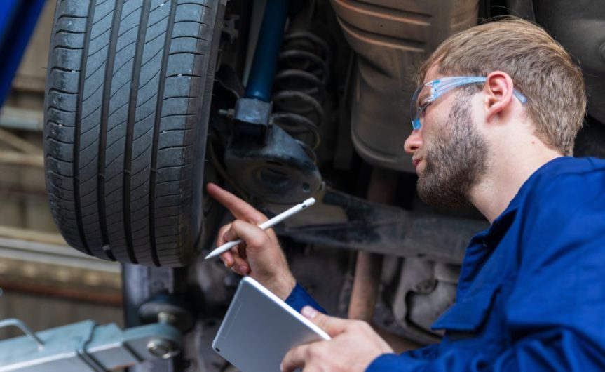 A man in a blue jumpsuit holding an electronic tablet and stylus examines a tire elevated on a hydraulic lift.