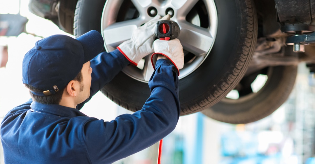A man in a blue work suit with a blue hat and white gloves loosens the lug nuts on a tire on an elevated car.