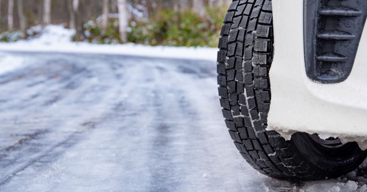A close-up of the winter tires of a white car on a road covered in ice with snowbanks and trees in the background.
