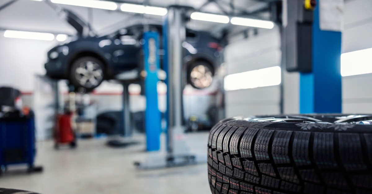 Two stacks of tires in an automotive repair shop. A dark gray car is raised on a large metal jack in the distance.
