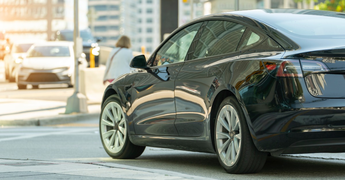 A black car parked on the street in a busy city during the daytime. The bright sun shines down onto the car and road.
