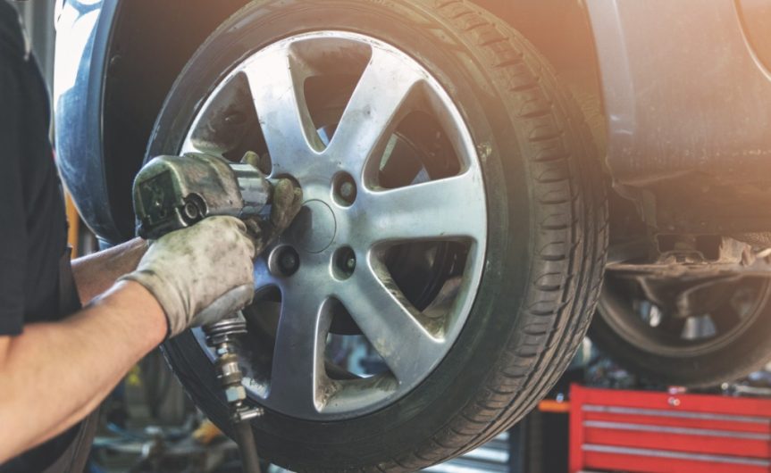 A close-up of a man using an impact wrench to remove the lug nuts from a wheel of a car on a lift in a repair shop.
