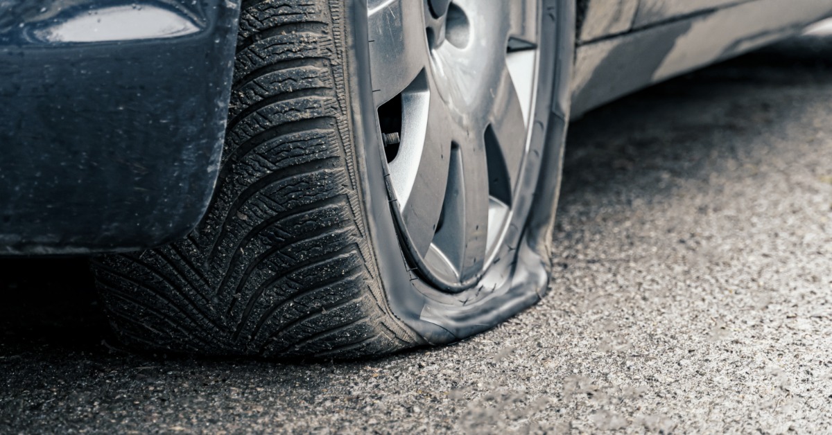 A black car parked on an asphalt road during the daytime. The driver’s side front tire on the car is flat.