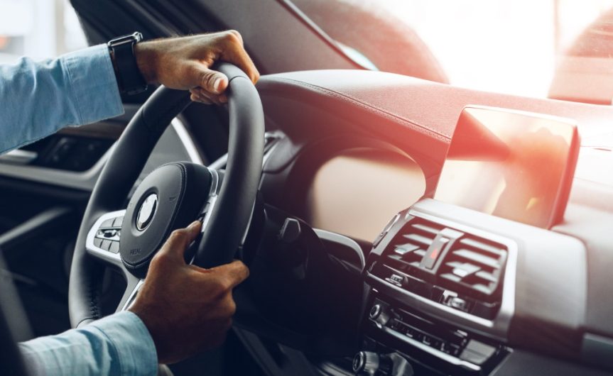 A person wearing a black watch drives a car on a bright day. The driver grasps the steering wheel with both hands.