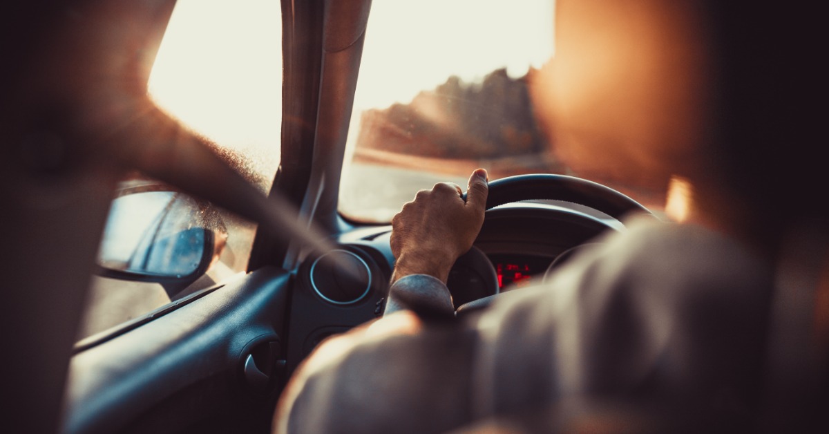 A person driving down a highway on a sunny day. The driver’s left hand sits on the car’s steering wheel.