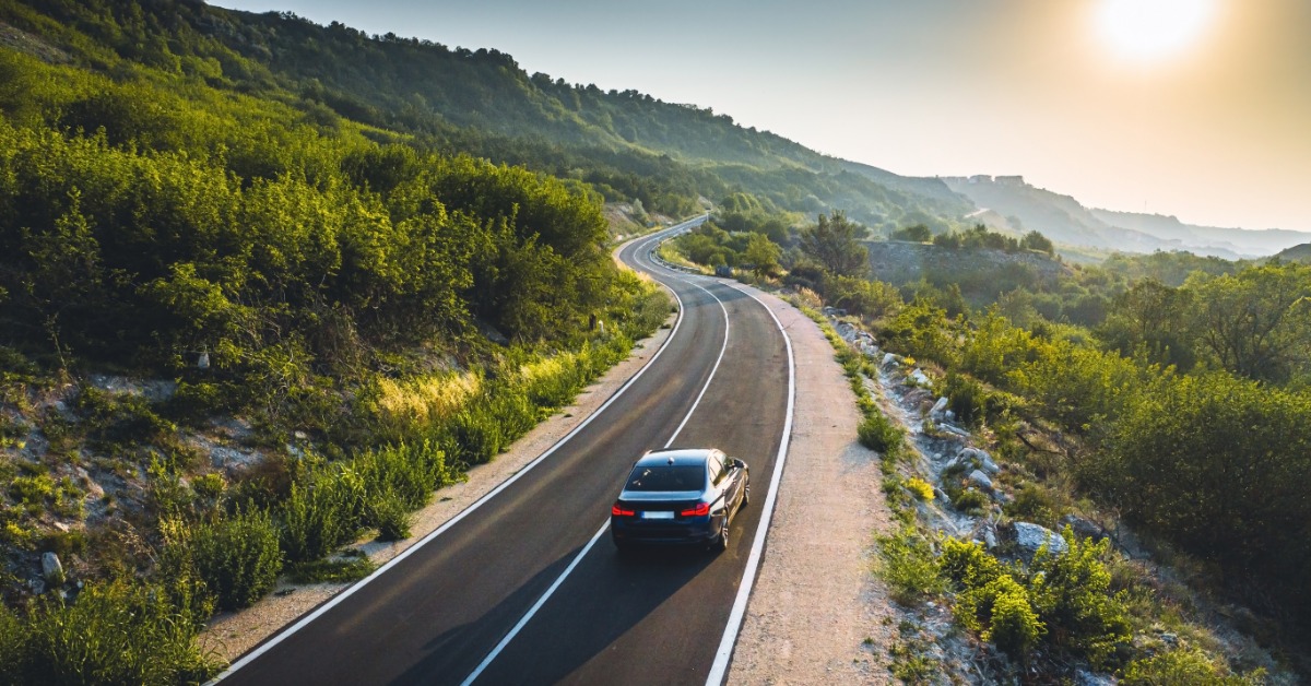 A black car drives down an empty highway on a bright, sunny day. Green trees grow next to the highway.