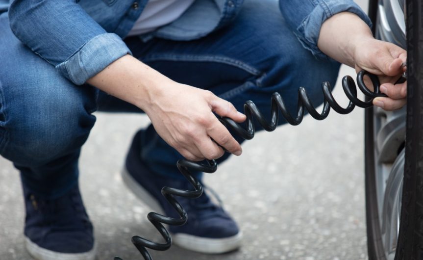 A person attaching a long, black hose to a car tire. The vehicle is parked outside during the daytime.
