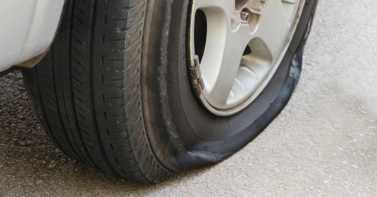 A clean gray car parked on a large patch of concrete in a brightly lit area. The car has a flat tire.