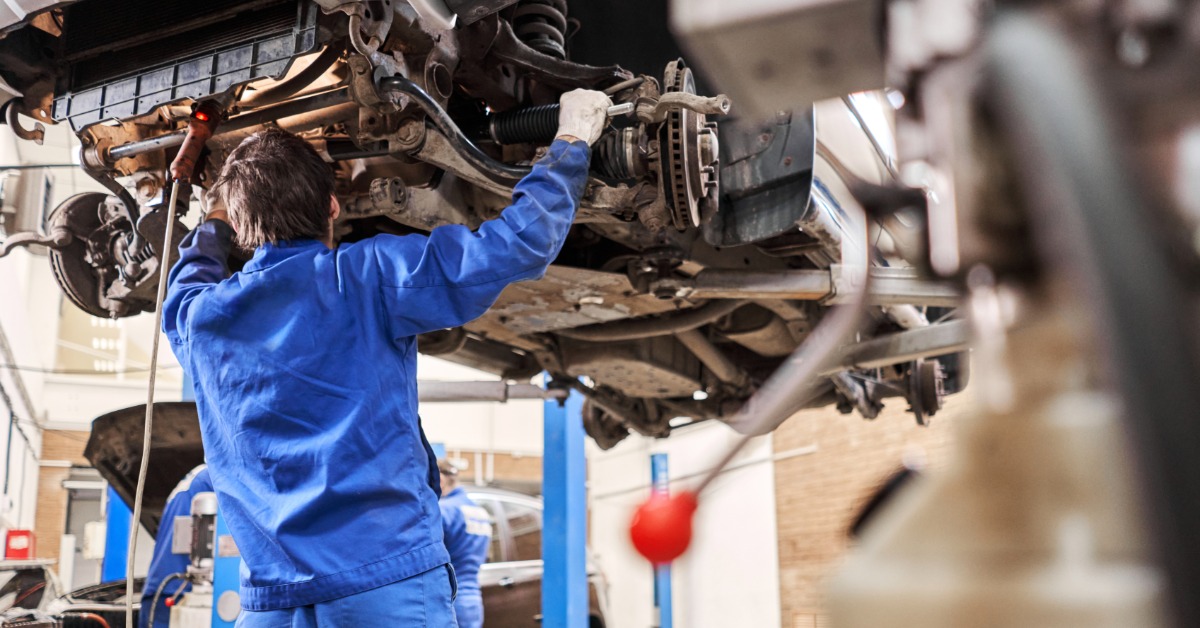 A rear view of a man in a white work suit working on the undercarriage of an elevated car on a hydraulic lift.